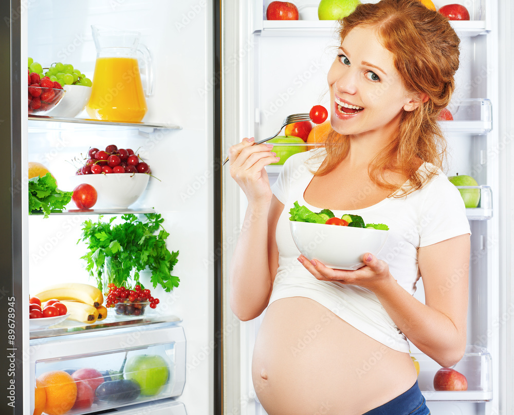 快乐的孕妇在冰箱附近吃沙拉