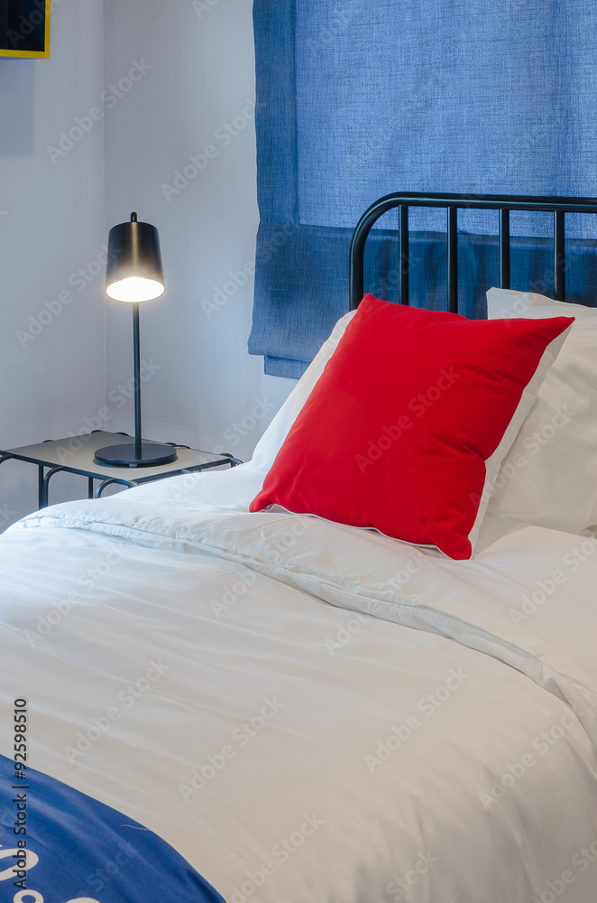 白色床上的红色枕头，蓝色窗帘，桌子上的黑色灯