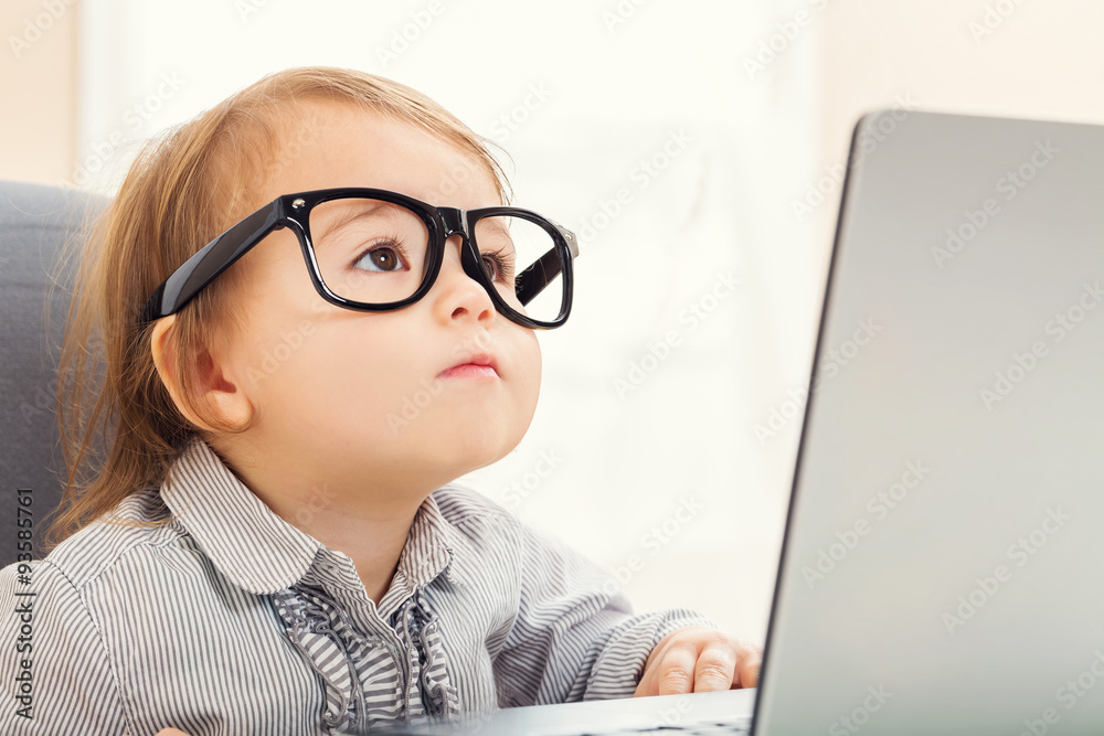 聪明的蹒跚学步的女孩在使用笔记本电脑时戴着大眼镜