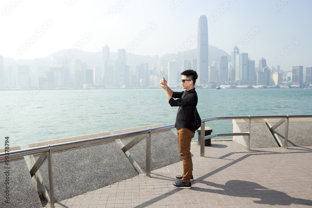 Fashionable young man taking photos with his smart phone in Victoria Harbor, Hong Kong
