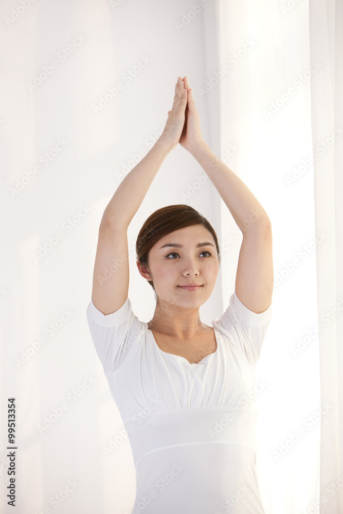 Young woman practicing yoga by a window with arms raised