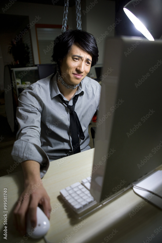一个穿着衬衫打着领带的年轻沮丧男子在使用电脑时被挂在隔间的链条上