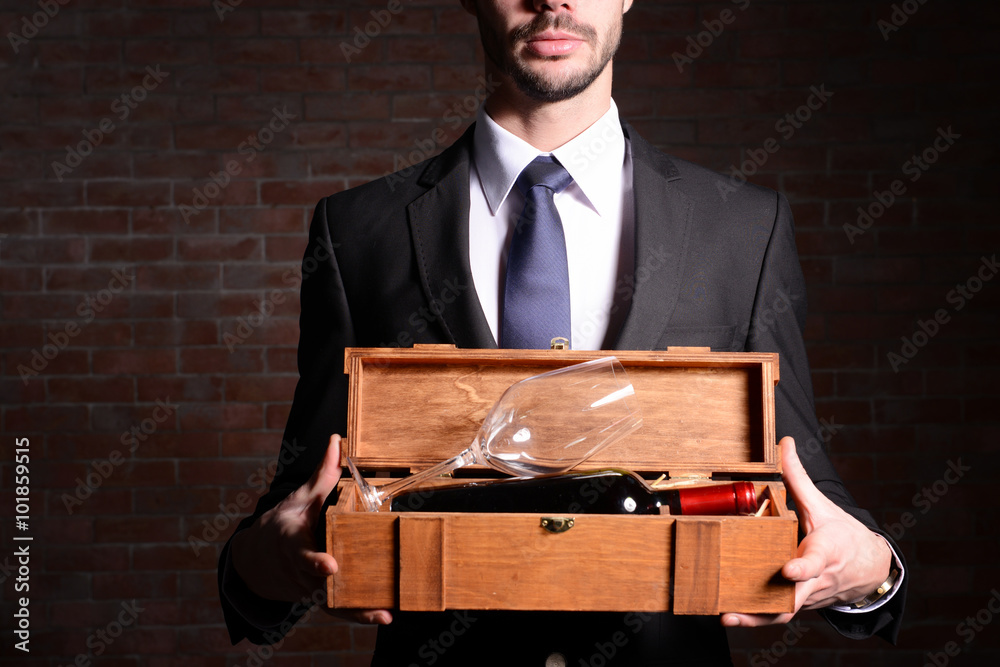砖墙背景上，一名男子拿着一瓶红酒和一个装在木箱里的玻璃