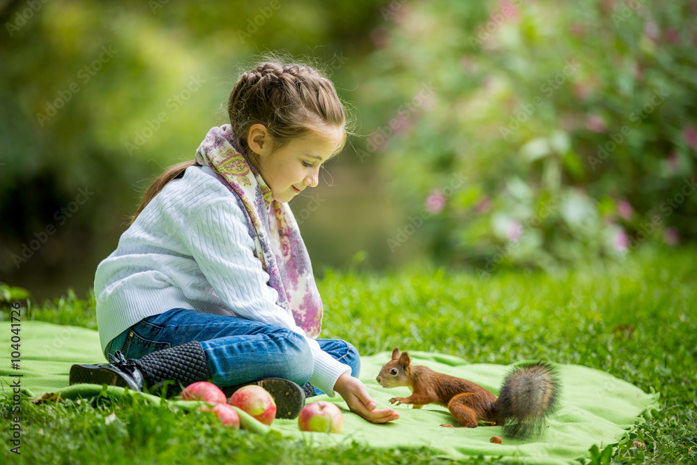 美丽的小女孩坐在绿色的草坪上，用坚果喂松鼠。