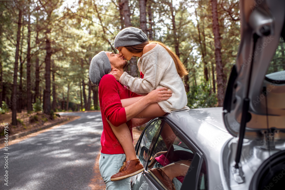 一对年轻可爱的情侣穿着运动衫，戴着帽子，在路边的汽车旁拥抱在一起，玩得很开心