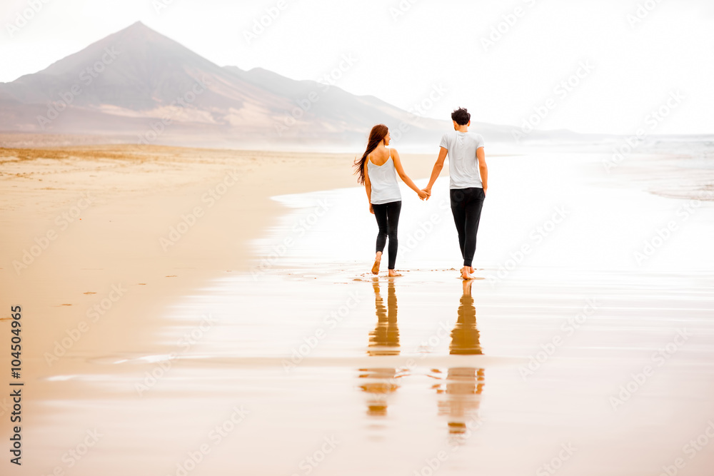 一对年轻可爱的情侣穿着白色t恤和黑色裤子，与mountai一起在美丽的沙滩上漫步