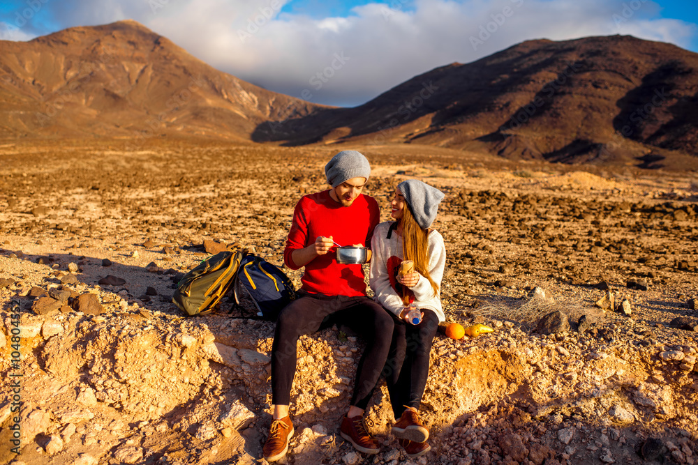 Young couple travelers eating healthy food from cooking pan sitting on the ground on desert mountain