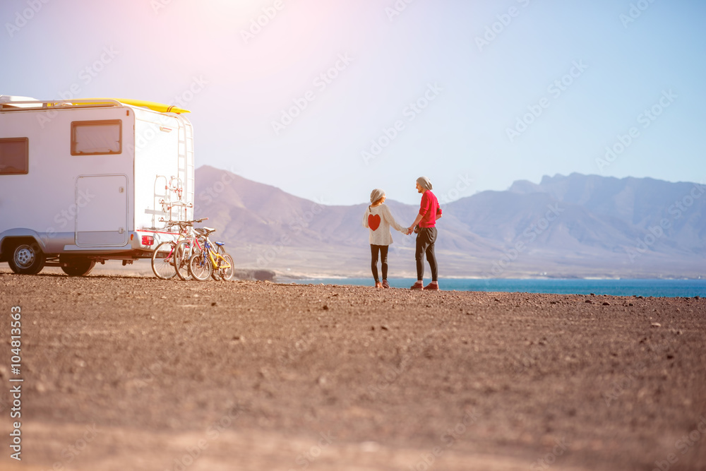 一对年轻夫妇站在荒凉的海边露营拖车附近，背景是群山