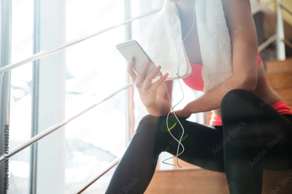 女运动员在健身房坐着使用带耳机的智能手机