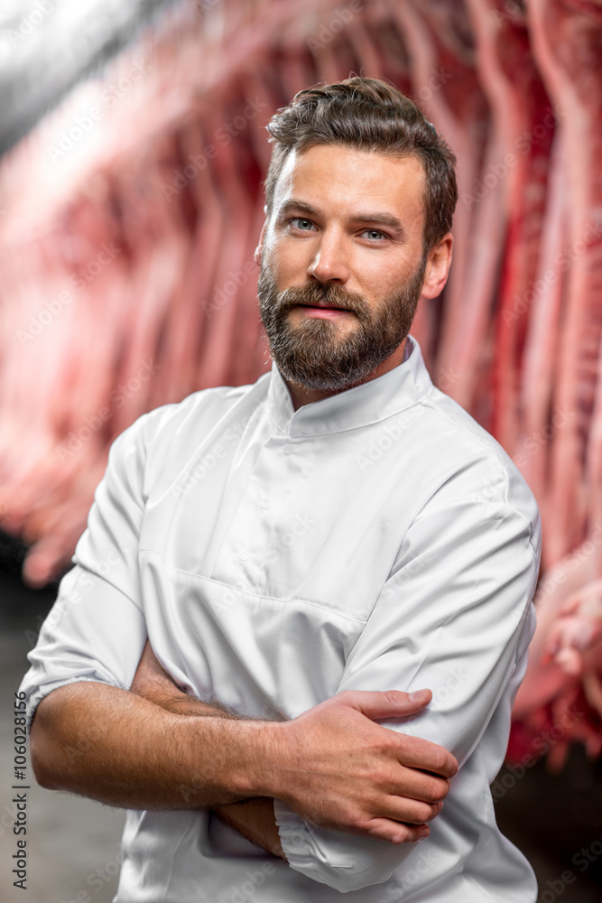 Portrait of a handsome butcher in white uniform at the meat manufacturing with pork carcasses on the