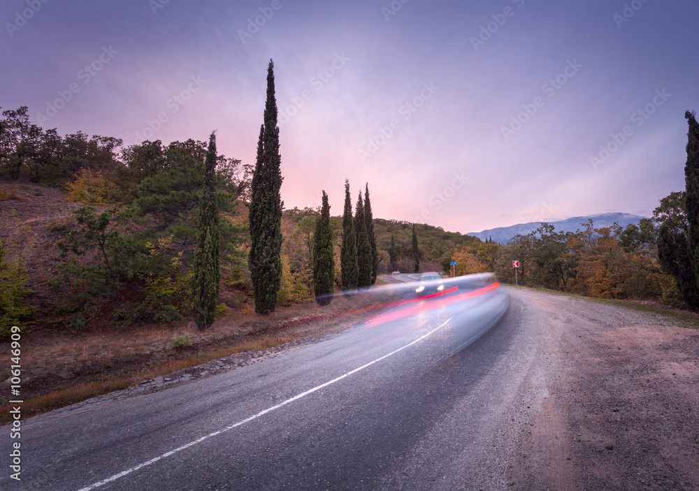 Mountain winding road passing through the forest with blurred car lights in motion with colorful sky