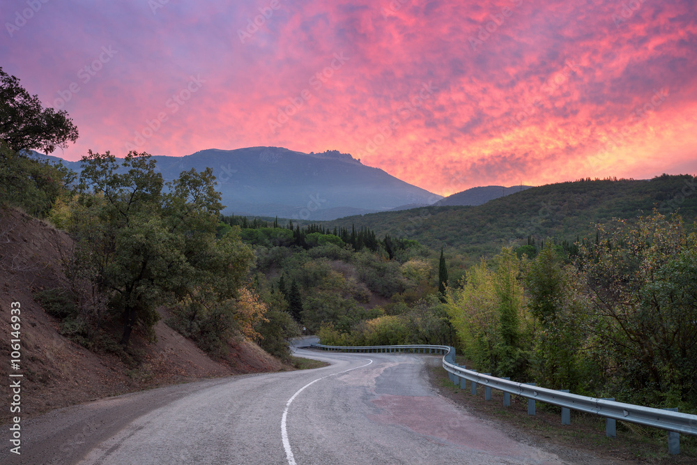 Mountain winding road passing through the forest with dramatic colorful sky and red clouds at sunset