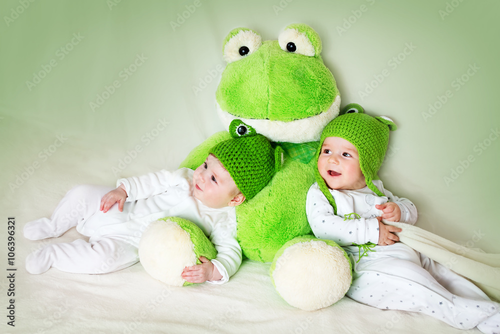 两个可爱的婴儿戴着青蛙帽躺着，手里拿着一个柔软的玩具