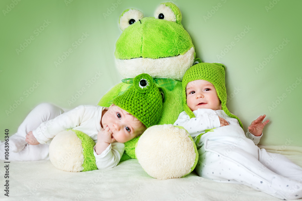 两个可爱的婴儿戴着青蛙帽躺着，手里拿着一个软玩具