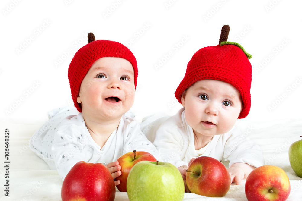 两个可爱的婴儿戴着帽子躺在柔软的毯子上，上面放着苹果