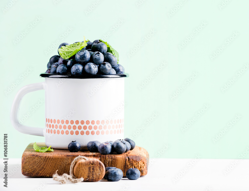 Fresh ripe blueberries in country style enamel mug on rustic wooden board over mint background