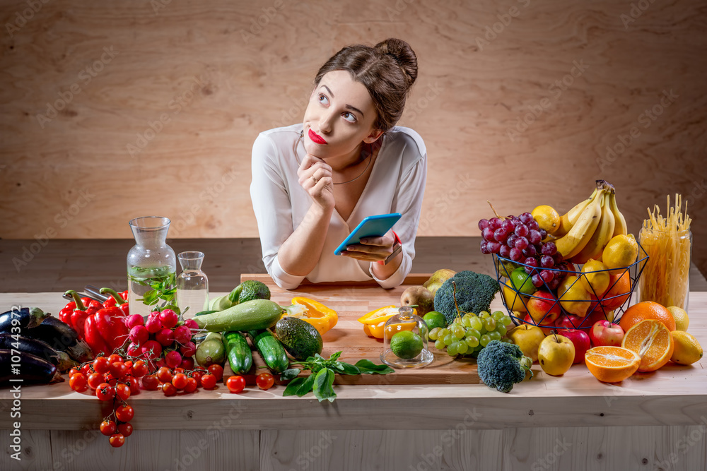 年轻漂亮的女人用智能手机坐在装满水果和蔬菜的桌子旁