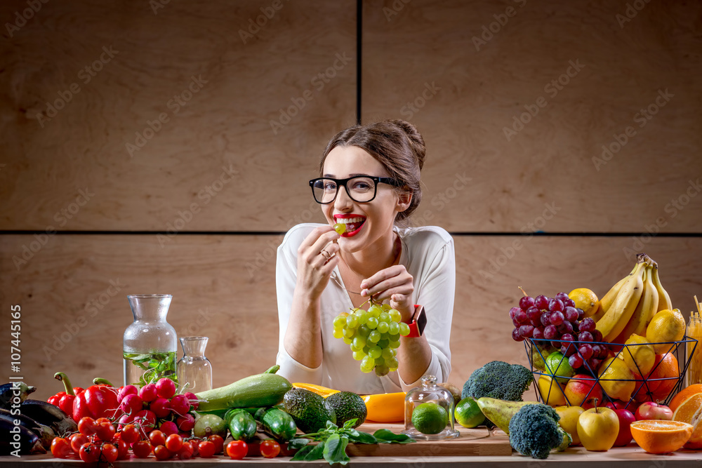 年轻可爱的女人在木制室内装满水果和蔬菜的桌子上吃葡萄