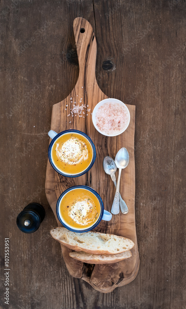 Homemade pumpkin cream soup in enamel mugs with herbs and fresh bread slices on olive serving board 