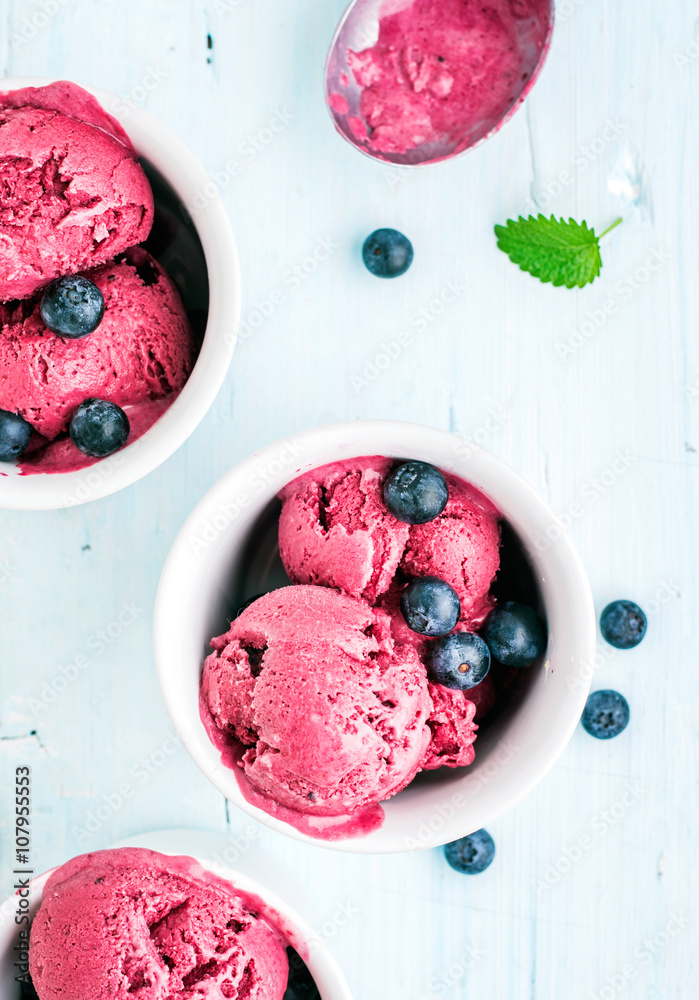 Homemade blueberry ice cream scoops with fresh berries and mint leaves in cups over light blue backg