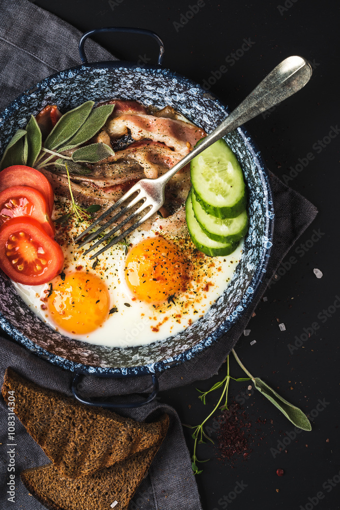 Breakfast set. Pan of fried eggs with bacon, fresh tomato, cucumber, sage and bread on dark serving 