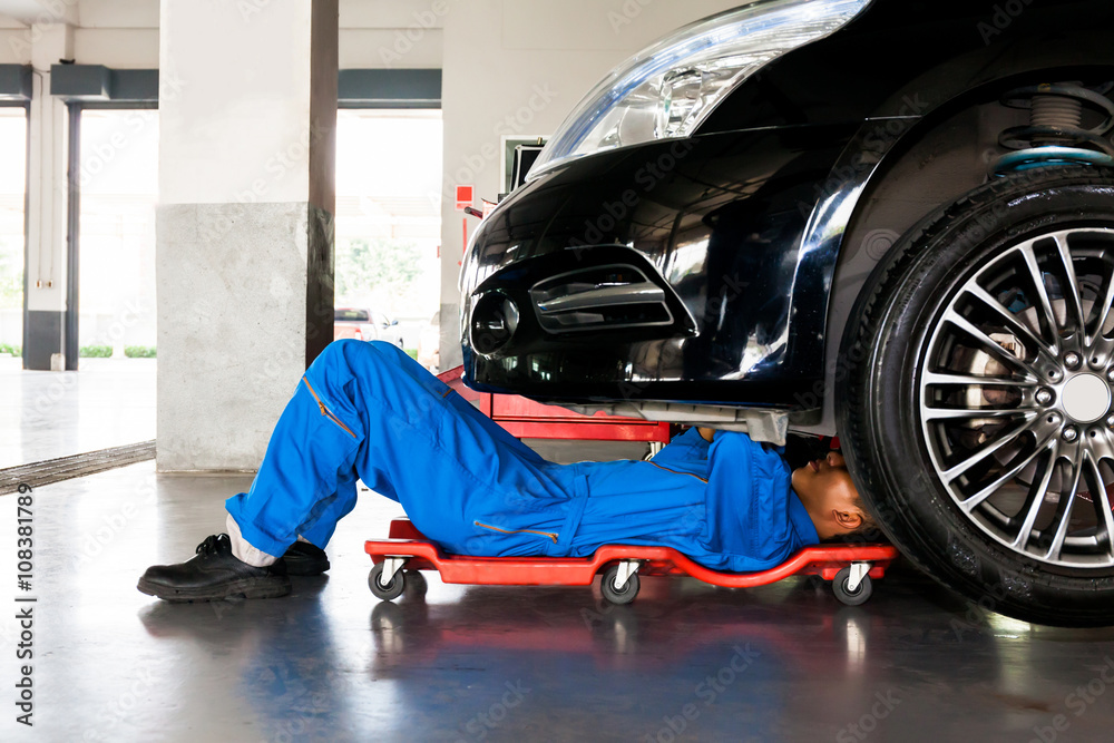 身穿蓝色制服的机修工躺在汽车维修库的车底工作