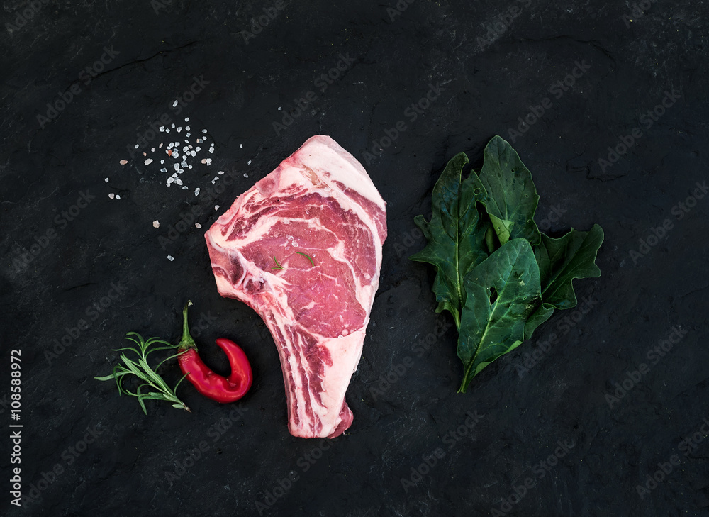 Raw fresh meat ribeye steak with salt, chili pepper, rosemary and spinach over black slate stone bac