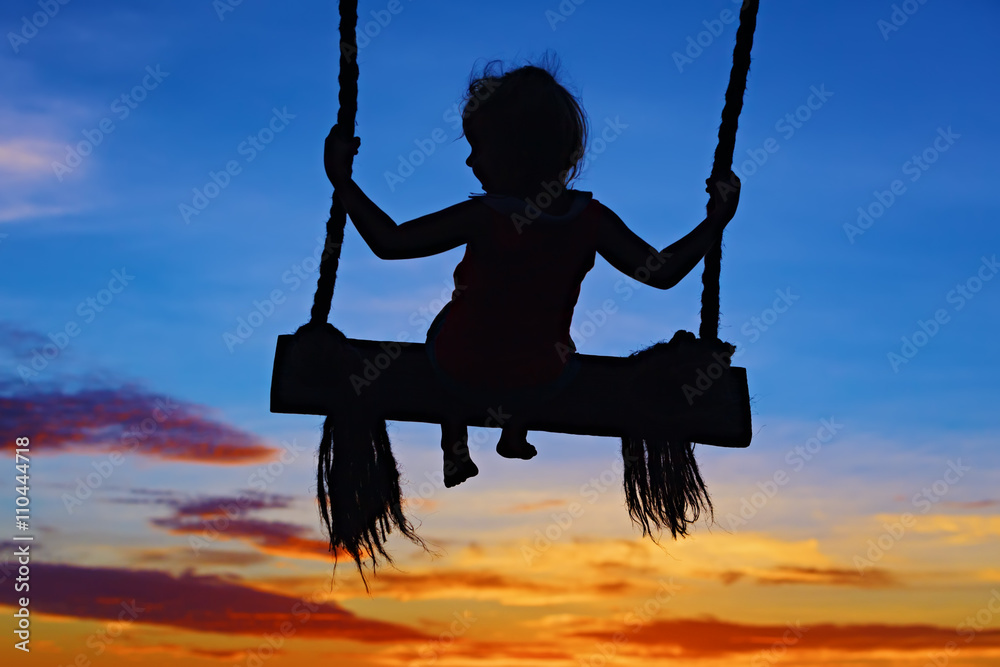 蓝橙色日落天空背景上，女婴在绳索秋千上快乐地高高飞翔的黑色剪影