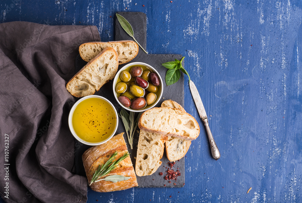 地中海小吃套装。黑石板石板上的橄榄、油、香草和切好的ciabatta面包