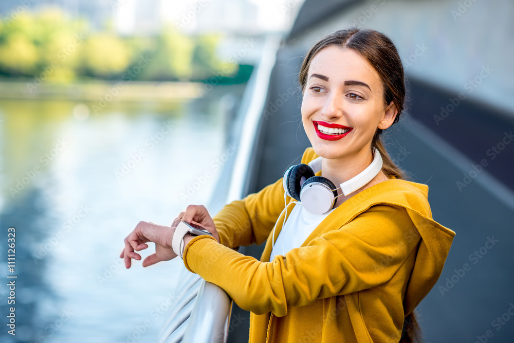 穿着黄色毛衣的年轻运动女性在摩天大楼林立的现代桥上看着智能手表