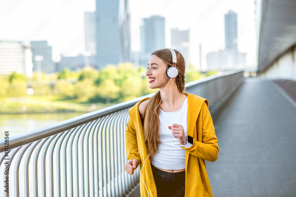 戴着耳机，穿着黄色毛衣的年轻运动女性在摩天大楼林立的现代桥上奔跑