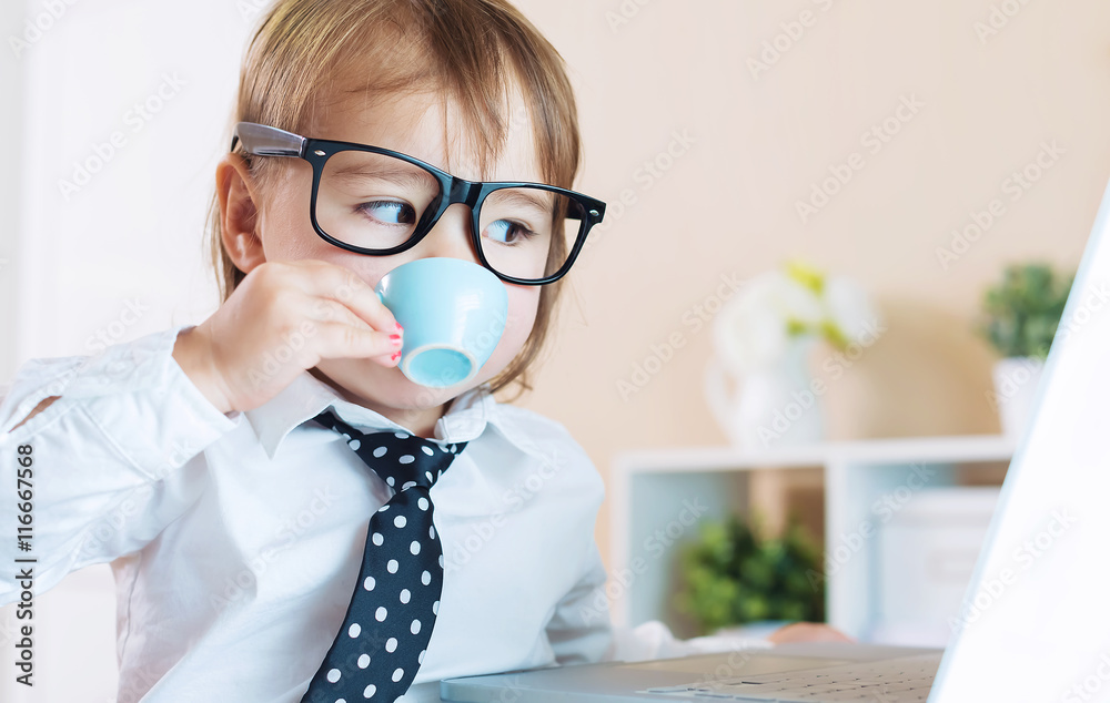 戴着眼镜的聪明蹒跚学步的女孩一边用笔记本电脑一边喝咖啡