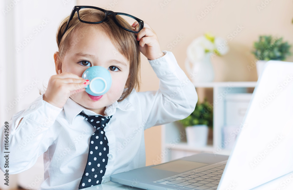 Smart toddler girl with glasses drinking coffee while using a laptop