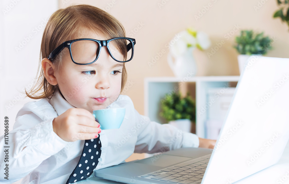 戴着眼镜的聪明蹒跚学步的女孩一边用笔记本电脑一边喝咖啡