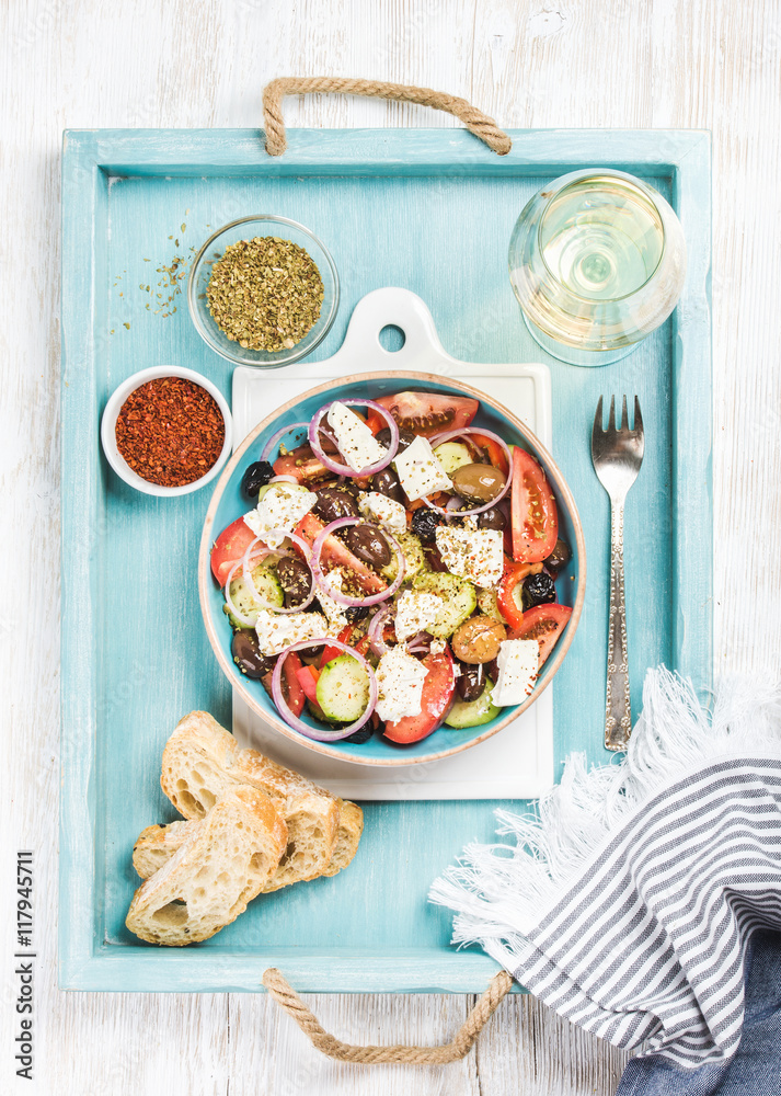 Greek salad with bread slices, oregano, pepper and glass of white wine on blue painted wooden tray o