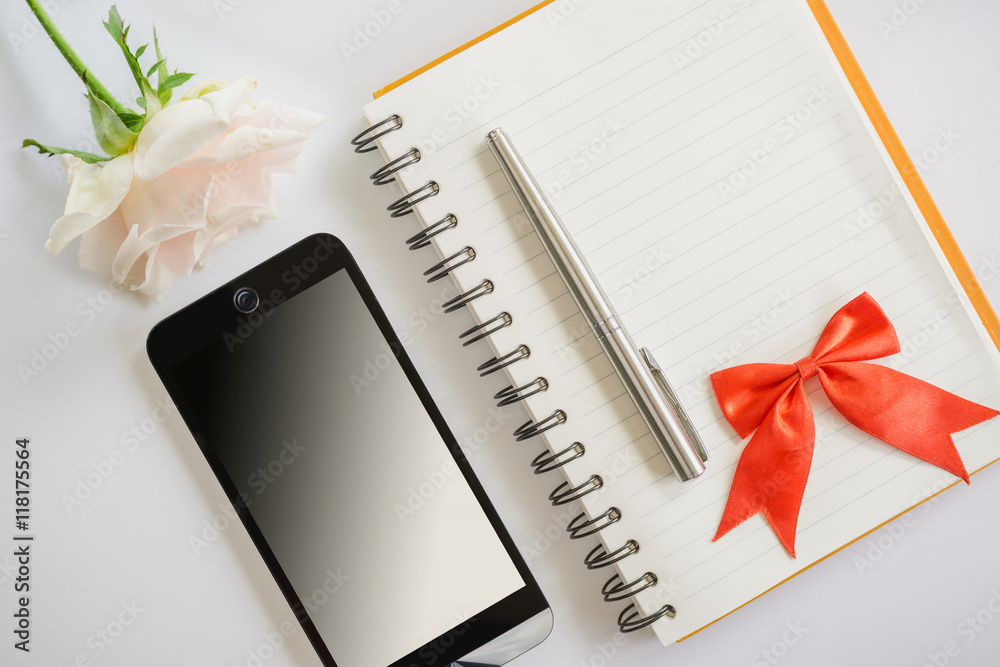 空白笔记本上的红色蝴蝶结和笔银，粉色玫瑰花和手机俯视图
