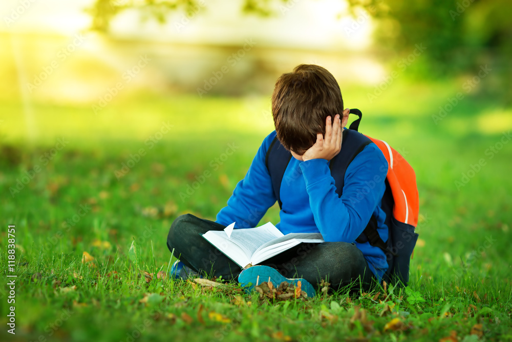 男孩背着帆布背包坐在公园里看书。孩子背着背包