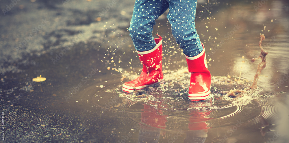 穿着红色橡胶靴的孩子的腿在秋天的水坑里跳跃