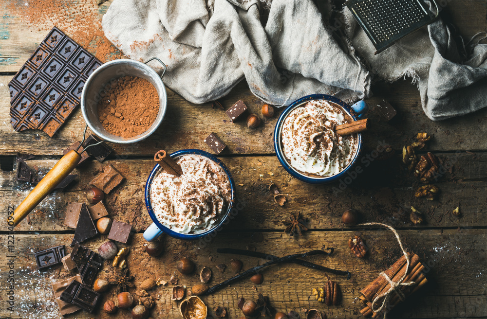 Hot chocolate with whipped cream, nuts and cinnamon in enamel mugs with ingredients around on rustic