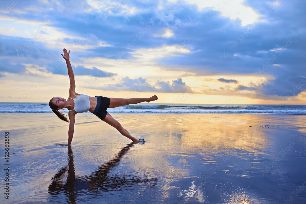 活跃的运动型女性在黑沙滩上摆瑜伽姿势，保持健康。日落蓝海