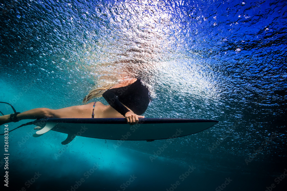 穿着比基尼的苗条金发女孩，一个冲浪者，带着冲浪板，鸭子在海水反射下潜水