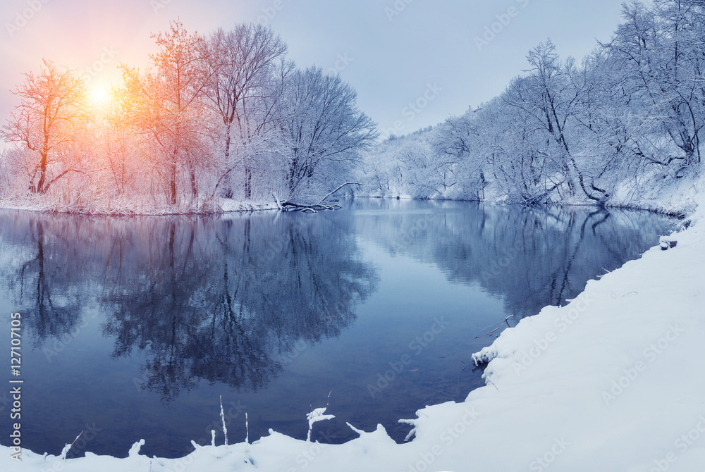 日落时河边的冬季森林。白雪皑皑的树木、阳光和美丽的冰冻ri的全景景观