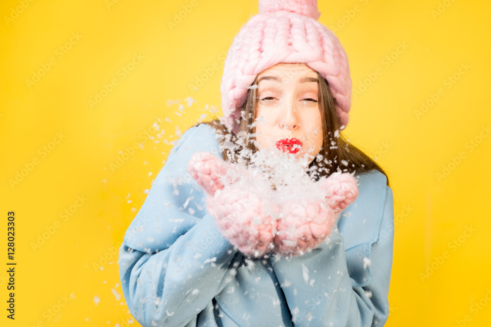 Young woman with knitted hat and gloves blowing snow standing on the yellow wall background. Colorfu