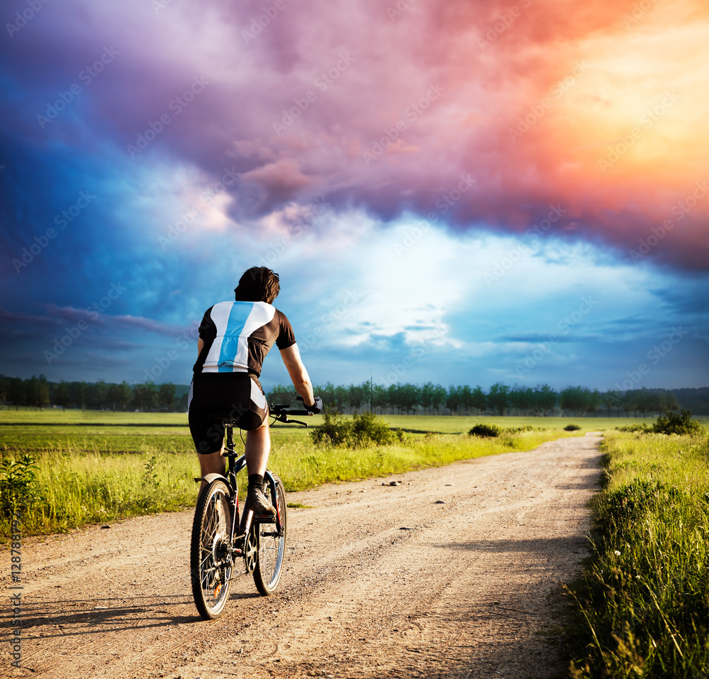 暴风雨日落时，一名男子在乡村公路上骑自行车。骑自行车的人的后视。戏剧性的晚云和S