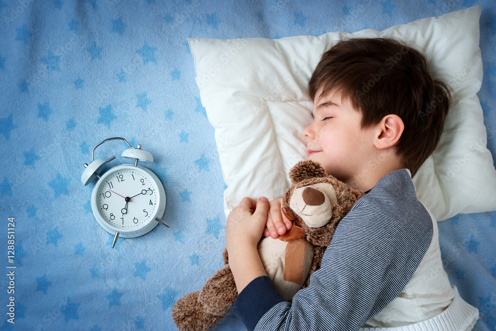 六岁的孩子睡在床上，枕头上有闹钟和泰迪熊