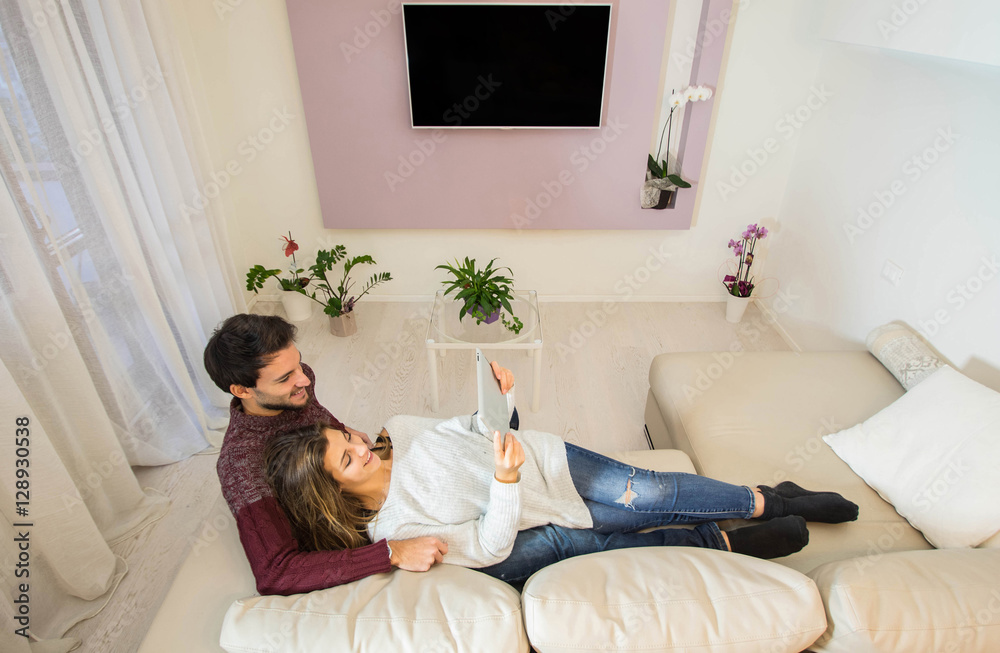 一对年轻夫妇躺在沙发上，在家里用平板电脑玩得很开心。