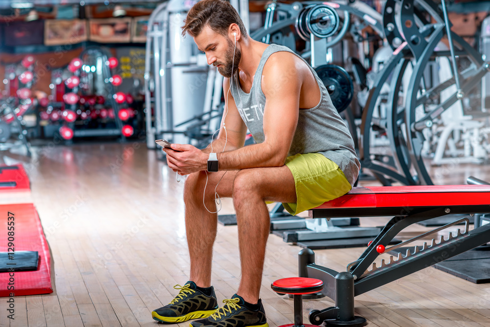 Muscular man using mobile phone sitting on the bench after the training in the gym