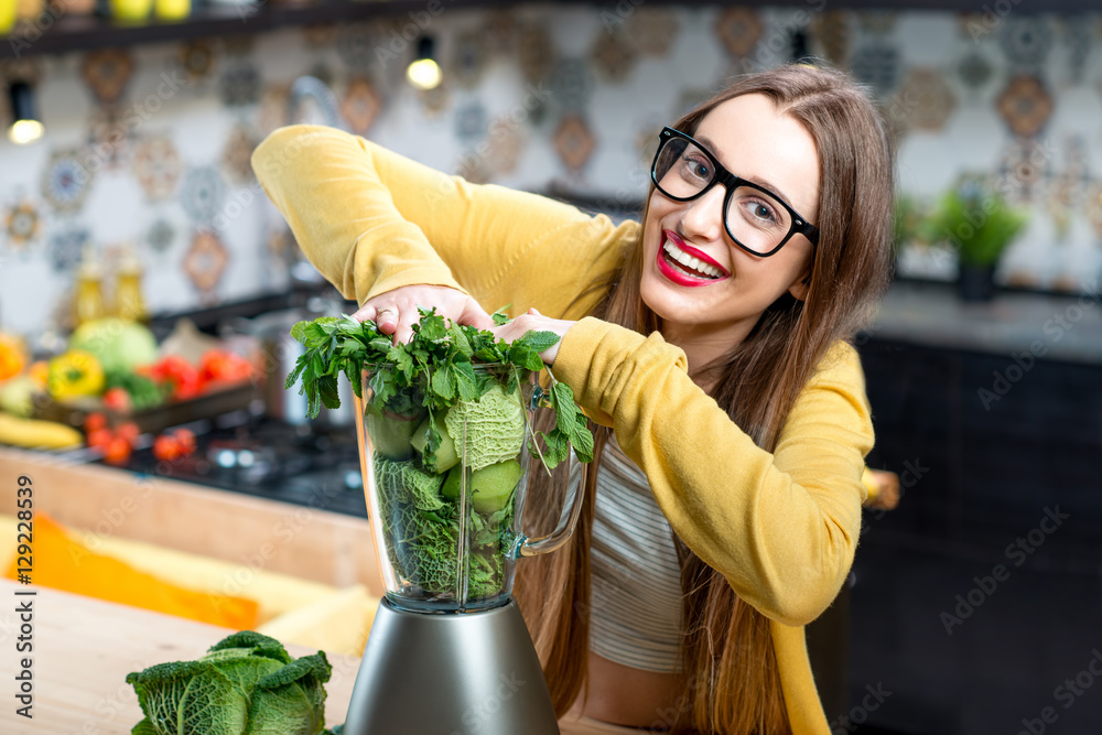 Young smiling woman pushing fresh greens into the blender in the kitchen at home. Healthy vegetarian