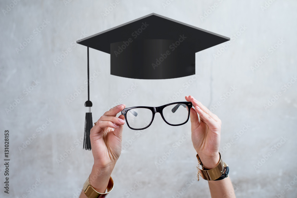 Female hands holding eyeglasses on the gray wall background with graffic academic hat