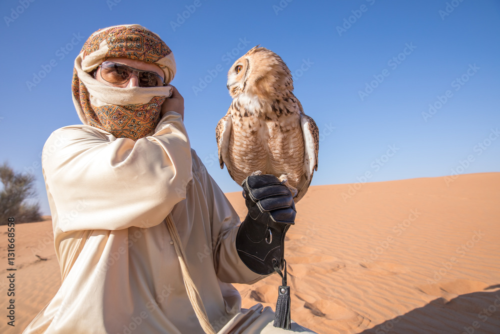 年轻的雄性法老鹰鸮（bubo ascalaphus）与一只穿着传统服饰的阿拉伯雄性在沙漠中